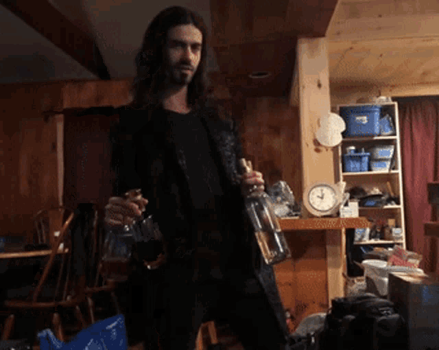a man with long hair is holding a bottle in a room with a clock on the wall