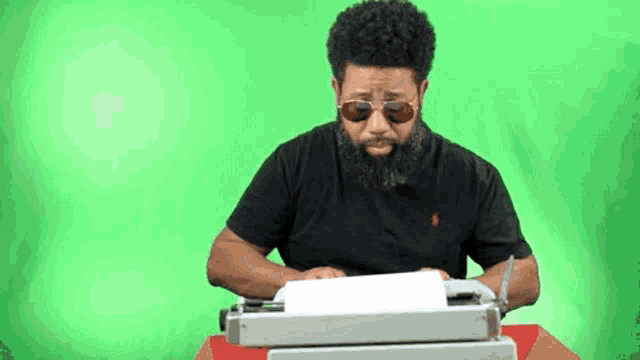a man with a beard is typing on a typewriter while wearing sunglasses .