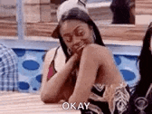 a woman is sitting at a table with her hands on her chin and a caption that says `` okay '' .