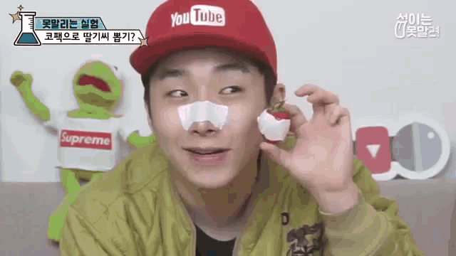 a man wearing a supreme shirt and a youtube hat is holding a strawberry in his hand .