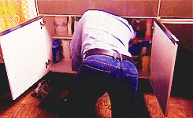 a man in a blue shirt is looking under a sink in a kitchen