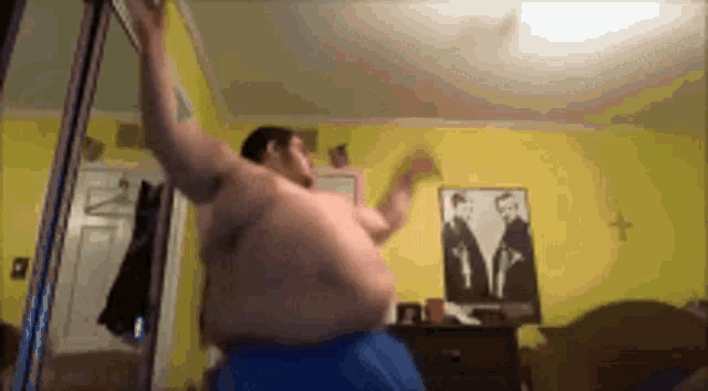 a shirtless man is dancing in a bedroom with a picture of two men on the wall