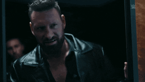 a man in a black leather jacket stands in a dark room with a w logo on the wall
