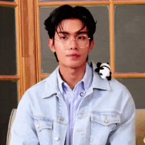 a young man wearing glasses and a denim jacket is sitting in front of a window with a stuffed panda .
