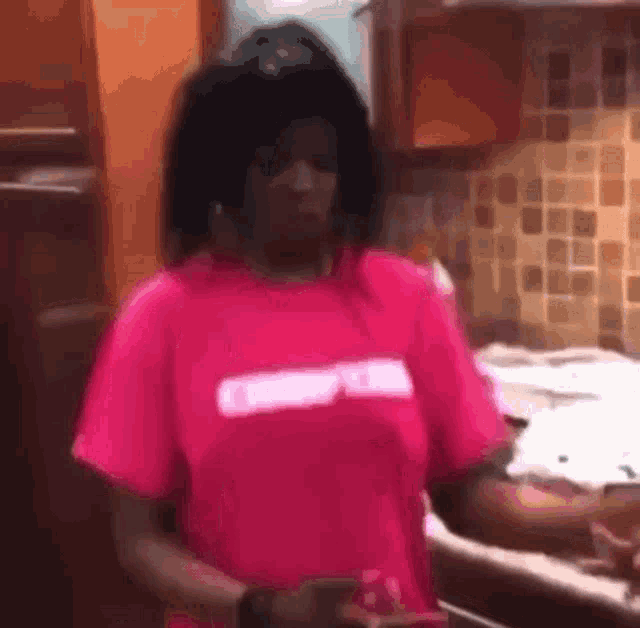 a woman in a pink t-shirt is standing in a kitchen next to a stove .