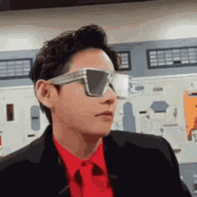 a man wearing sunglasses and a red shirt and tie is standing in a room .