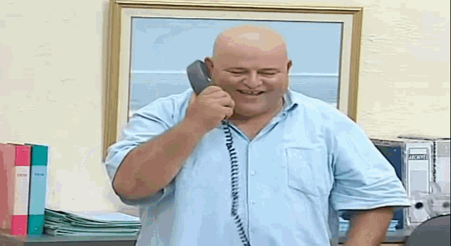 a bald man is talking on a telephone with binders in the background .