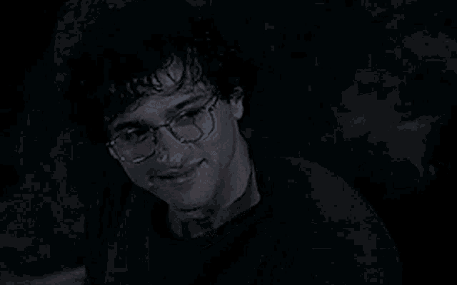 a man wearing glasses and a black shirt is smiling in a dark room .