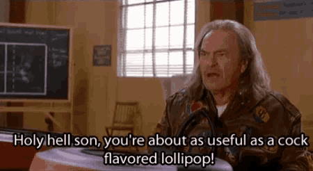 a man with long hair is sitting at a table with a lollipop in his hand and talking to another man .