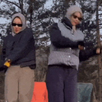 two people are standing next to each other in the woods wearing sunglasses and jackets .