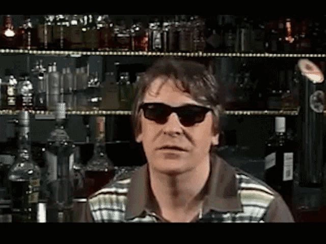 a man wearing sunglasses is standing in front of a bar filled with alcohol bottles .