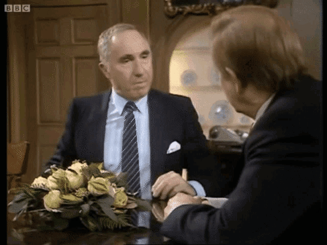 two men are sitting at a table with a bouquet of flowers in front of them and the bbc logo is visible