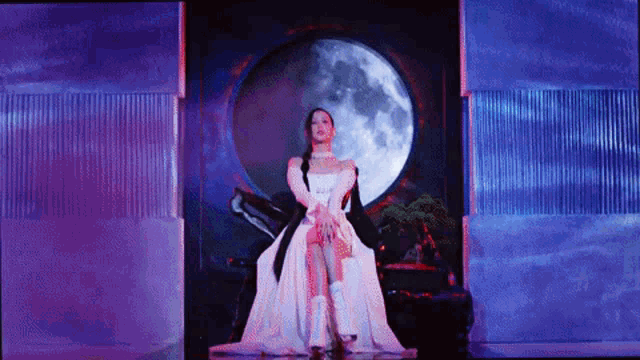 a woman in a long white dress is standing in front of a full moon on a stage .