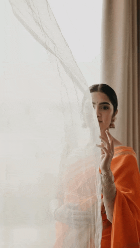 a woman in an orange saree is standing in front of a window and smiling