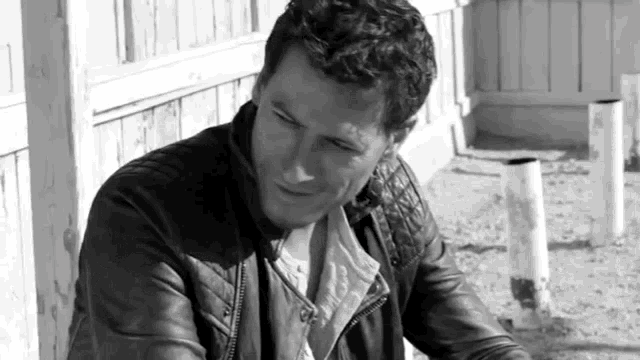 a man in a leather jacket is sitting on the ground in front of a fence .