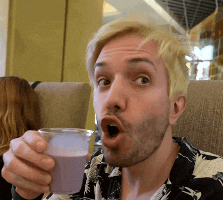 a man with blonde hair and a beard is holding a cup of purple liquid with his mouth open