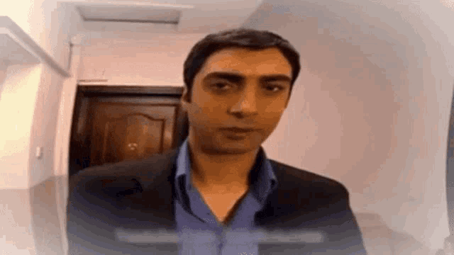 a man in a suit and blue shirt is standing in front of a door looking at the camera .
