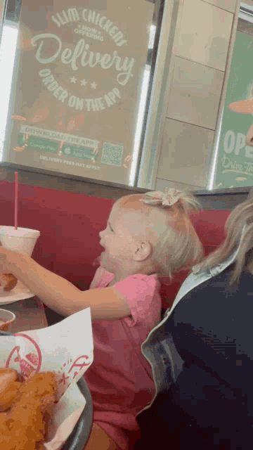 a little girl is sitting at a table in front of a sign that says slim chickens delivery
