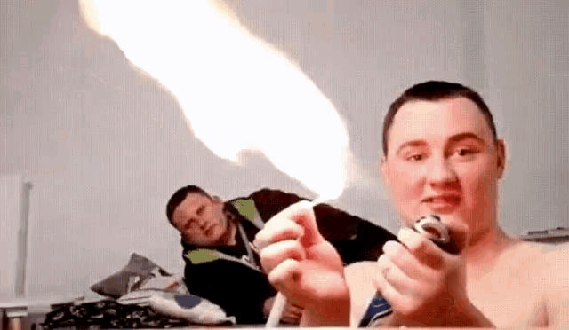 a man is holding a lighter with a flame coming out of it and another man is laying on a bed behind him