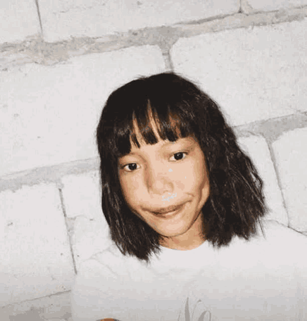 a young girl with short hair and bangs is sitting in front of a brick wall .