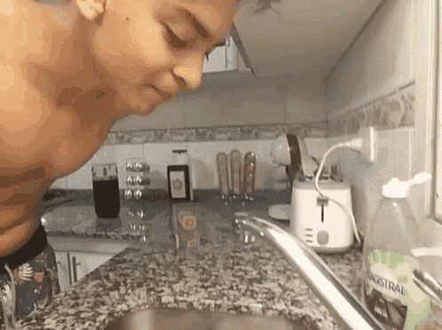 a man is washing his face in a kitchen sink with a bottle of magistral soap on the counter