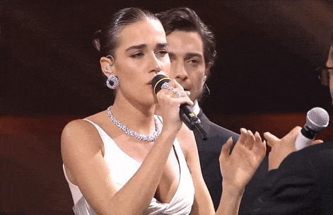 a woman in a white dress singing into a microphone with a man clapping behind her