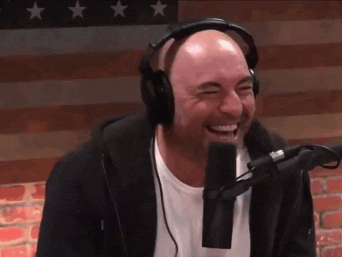 a bald man wearing headphones is laughing into a microphone while sitting in front of an american flag .