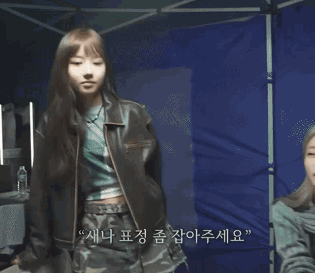 a woman in a leather jacket is standing in front of a blue wall with korean writing on it