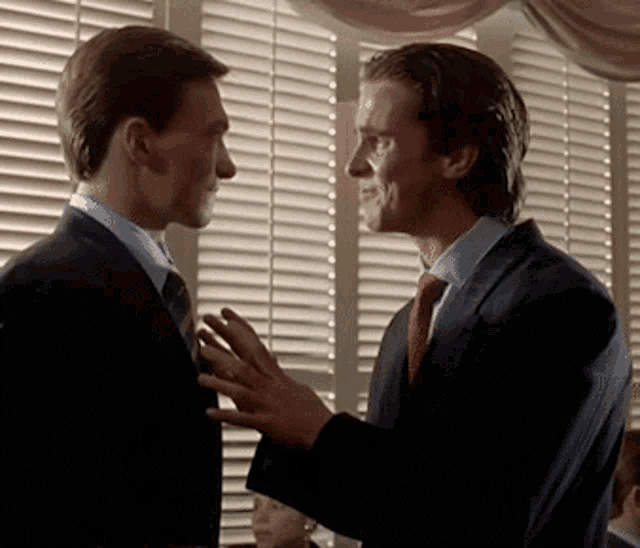 two men in suits and ties are talking to each other in front of a window with blinds .