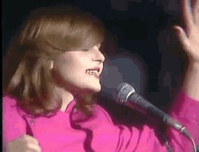 a woman in a pink sweater is singing into a microphone with her hands in the air .