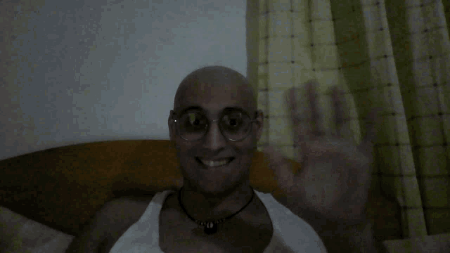 a man with glasses and a necklace is smiling and waving his hand