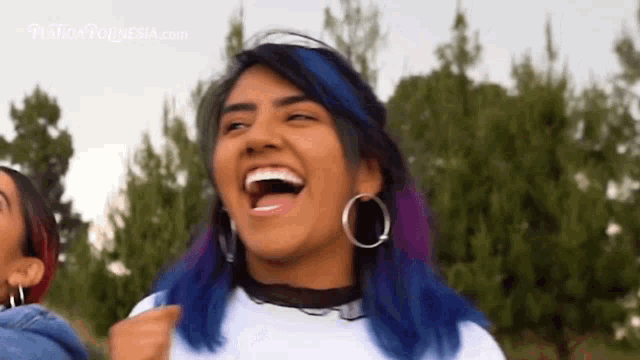 a woman with blue and purple hair and hoop earrings is laughing with her mouth open .