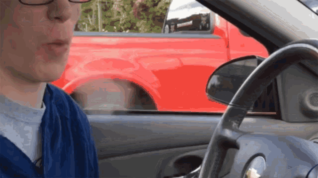 a man wearing glasses is driving a car with a red truck behind him