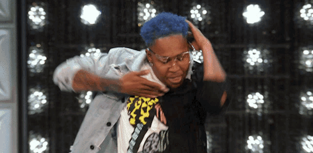 a person with blue hair and glasses holds their chest