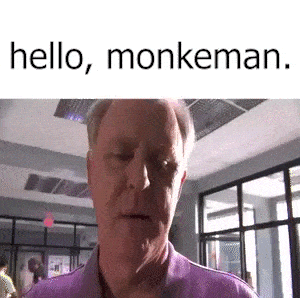 a man in a purple shirt is standing in a room with the words `` hello , monkeman '' written above him .