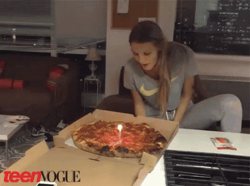 a woman blows out a candle on a pizza that is in a box that says teenvogue
