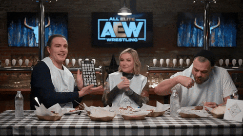 three people sit at a table in front of a aew wrestling sign
