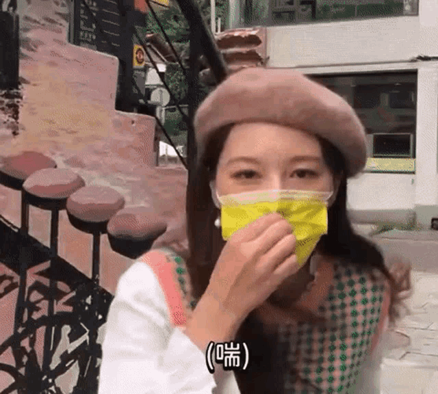 a woman wearing a beret and a yellow face mask is covering her mouth with her hand .