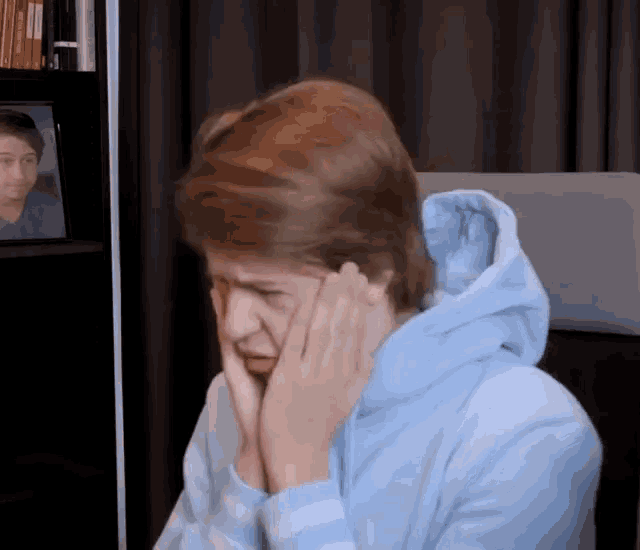 a man in a blue hoodie is covering his face with his hands .
