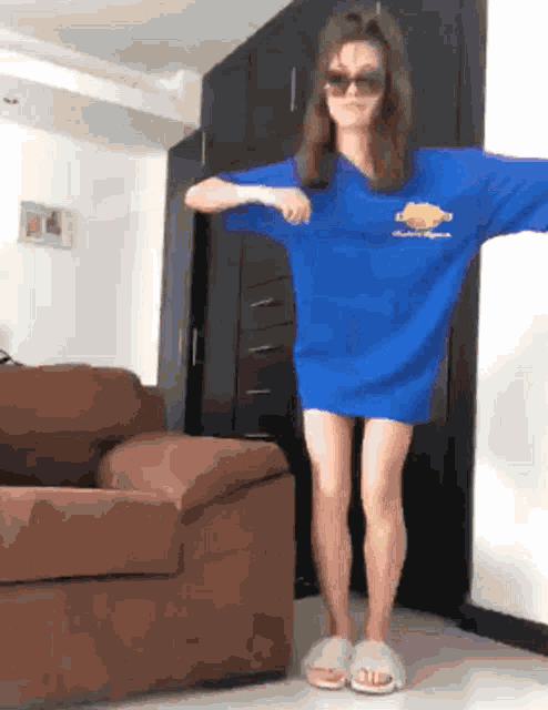 a woman wearing sunglasses and a blue shirt is standing in front of a brown couch