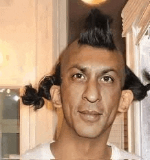 a man with a mohawk and pigtails on his head is standing in front of a window .