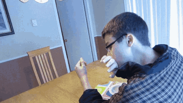a man wearing glasses sits at a table eating a piece of pizza