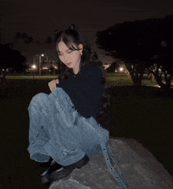 a girl in a black sweater and blue jeans sits on a rock