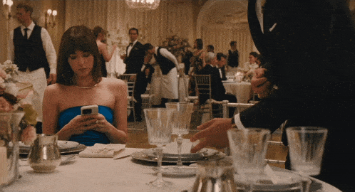 a woman in a blue dress sits at a table looking at her cell phone