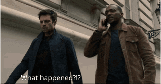 a man talking on a cell phone while another man stands next to him and says " what happened "