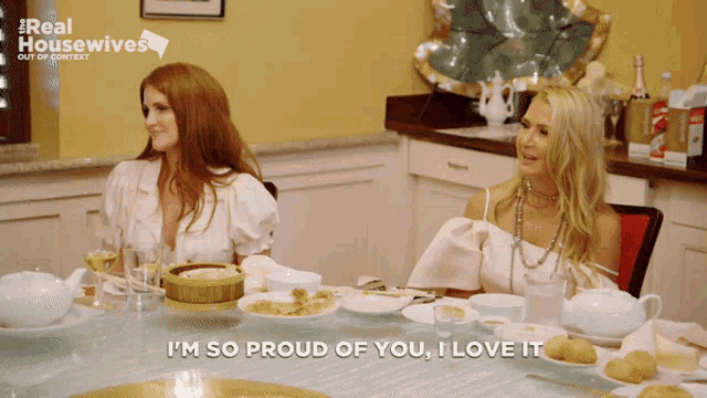 two women are sitting at a table with plates of food and one of them is saying i 'm so proud of you