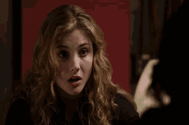 a woman with blonde curly hair is sitting in front of a red wall and making a funny face .