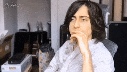 a man with long hair is sitting in a chair with his hand on his chin