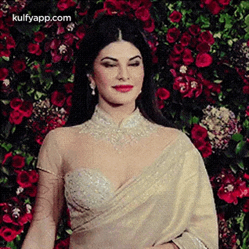 a woman in a white saree is standing in front of a wall of flowers .