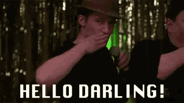a man in a hat is covering his face with his hand while holding a microphone and saying `` hello darling '' .